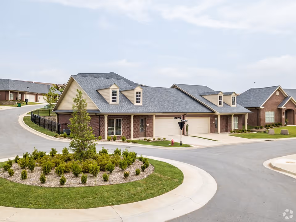 Exterior view of a residential-style building with brick walls and gray shingled roofs, surrounded by a curved road and landscaped greenery including a roundabout with shrubs and a small tree.