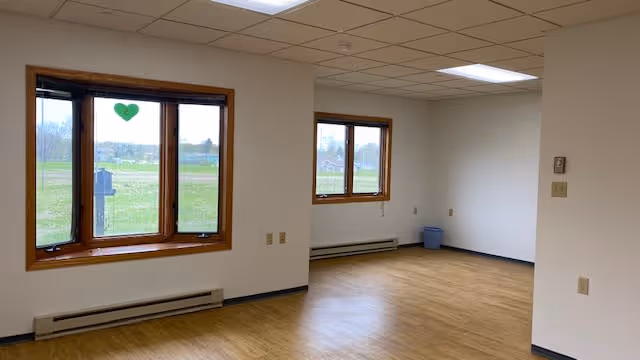 Empty room with light wood flooring, white walls, and two windows with wooden frames. One window has a green heart decoration. The room has a drop ceiling with fluorescent lights and baseboard heaters below the windows. A small blue trash bin is in the corner.