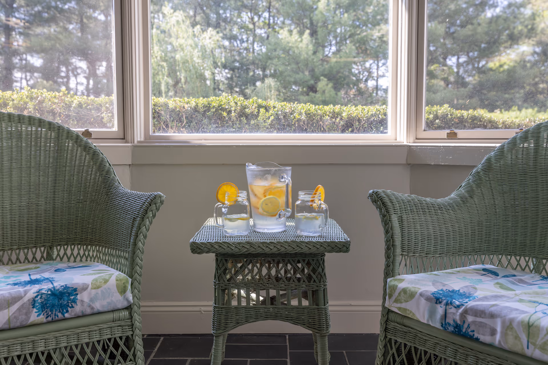 Two wicker armchairs flank a small wicker table holding a pitcher and glasses of lemon water in front of a large window overlooking hedges and trees.