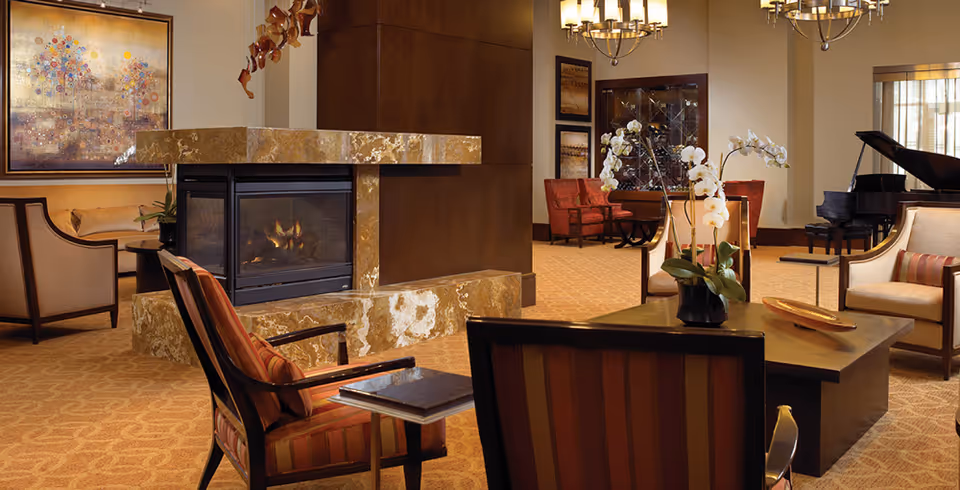 Elegant communal lounge with a central marble fireplace, upholstered seating, a coffee table with orchids, and a grand piano.