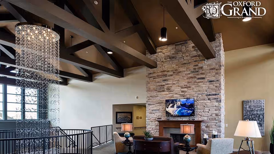 Spacious senior living lounge with exposed wood beams, a stone fireplace, seating areas, and a hanging crystal chandelier.