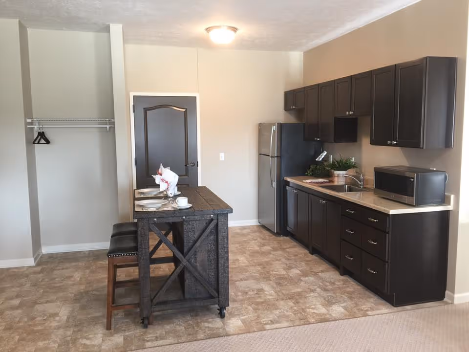 A small kitchen area with dark wood cabinets, a stainless steel refrigerator, a microwave on the countertop, and a sink. In front of the kitchen is a wooden island with two stools and place settings for two. To the left, there is an open closet space with a hanging rod and two hangers. The floor is tiled and the walls are painted beige.