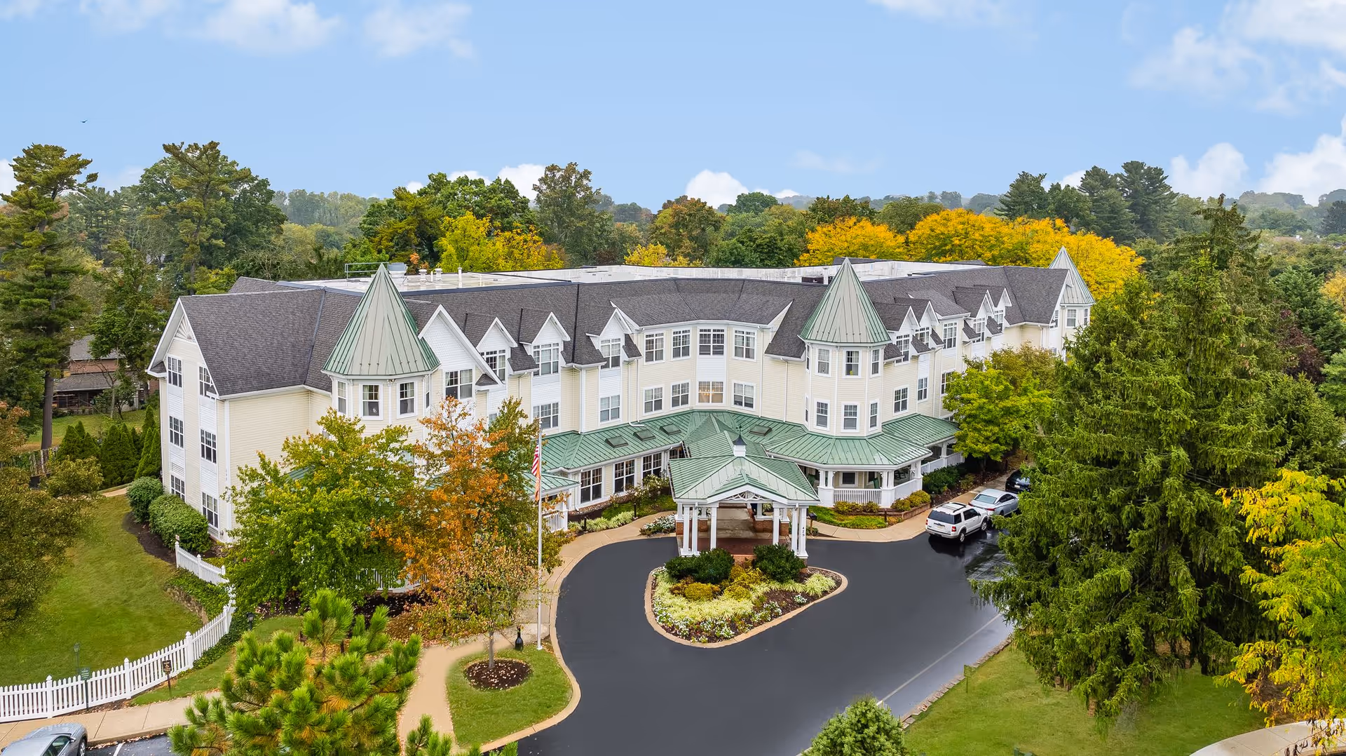 Aerial view of a large senior living facility building with white exterior walls and green roofs, surrounded by trees with autumn foliage. The building has multiple peaked roof sections and a covered entrance driveway with cars parked nearby. The facility is set in a lush, green landscape under a partly cloudy blue sky.