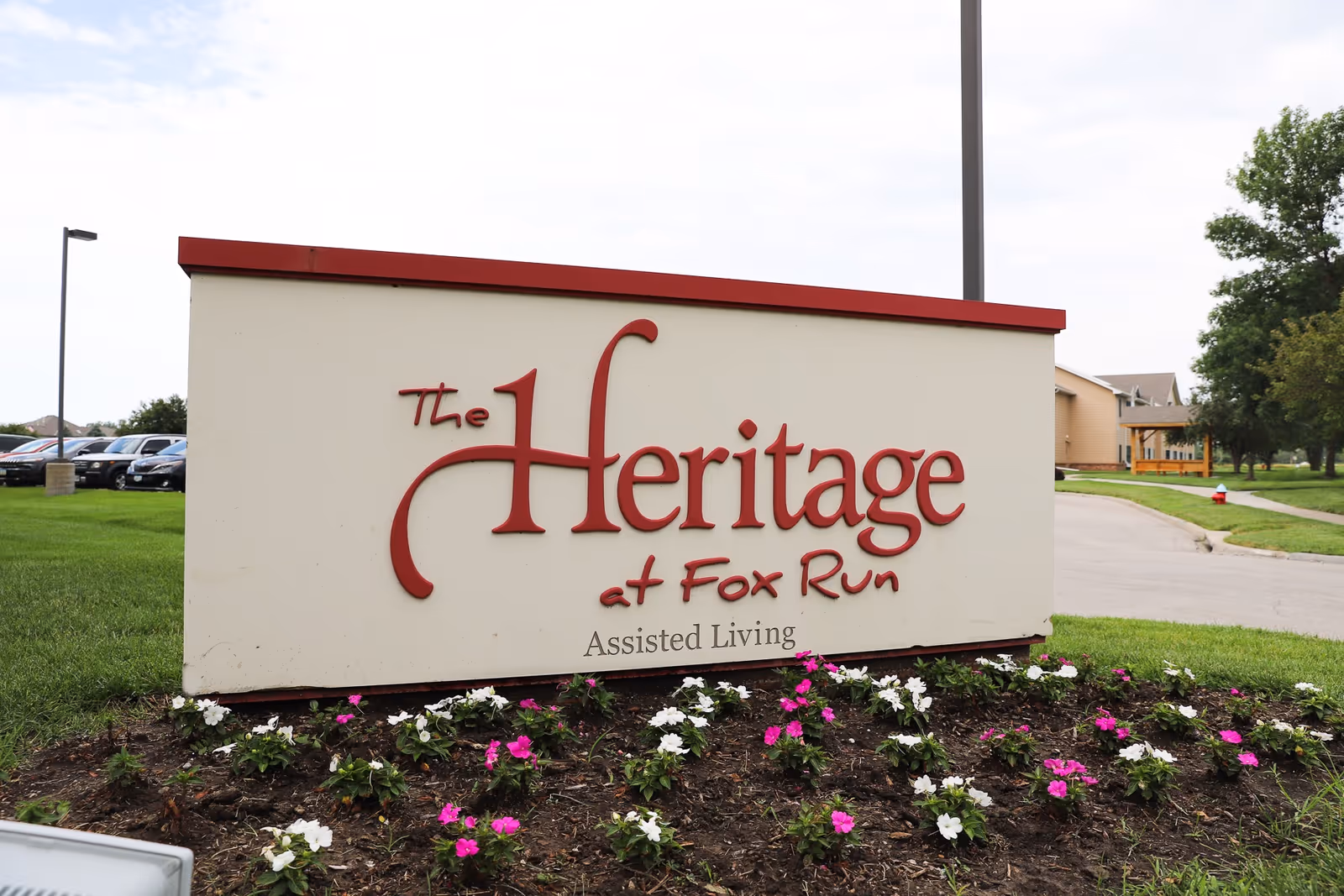 Outdoor sign for The Heritage at Fox Run Assisted Living facility with flowers planted in front and a parking lot and trees in the background.