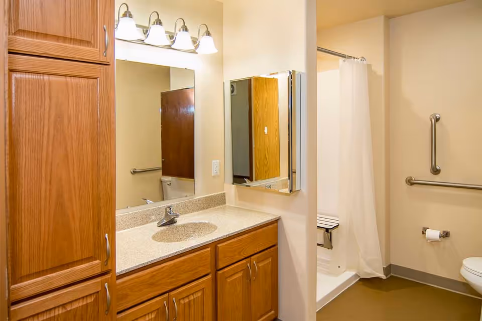 A bathroom with wooden cabinets and a beige countertop with a sink. Above the sink is a large mirror with four light fixtures. To the right, there is a shower area with a white curtain and a fold-down seat. The bathroom has grab bars near the toilet and shower for accessibility.
