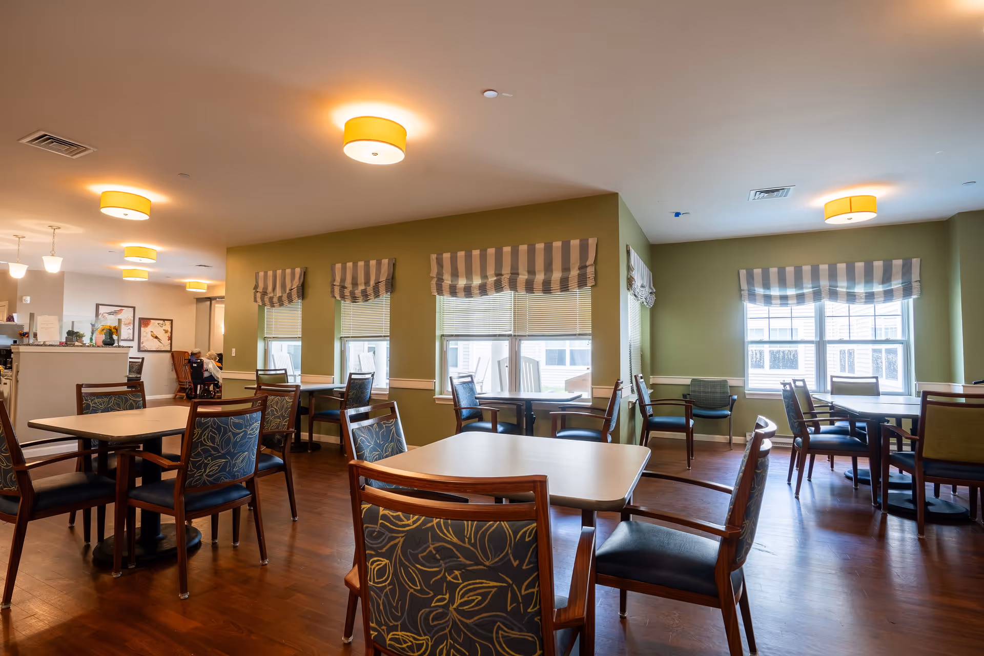 A dining room in an assisted living facility with multiple tables and chairs arranged neatly. The room has green walls, large windows with striped valances, and warm ceiling lights. There are a few residents seated in the background near the kitchen area.