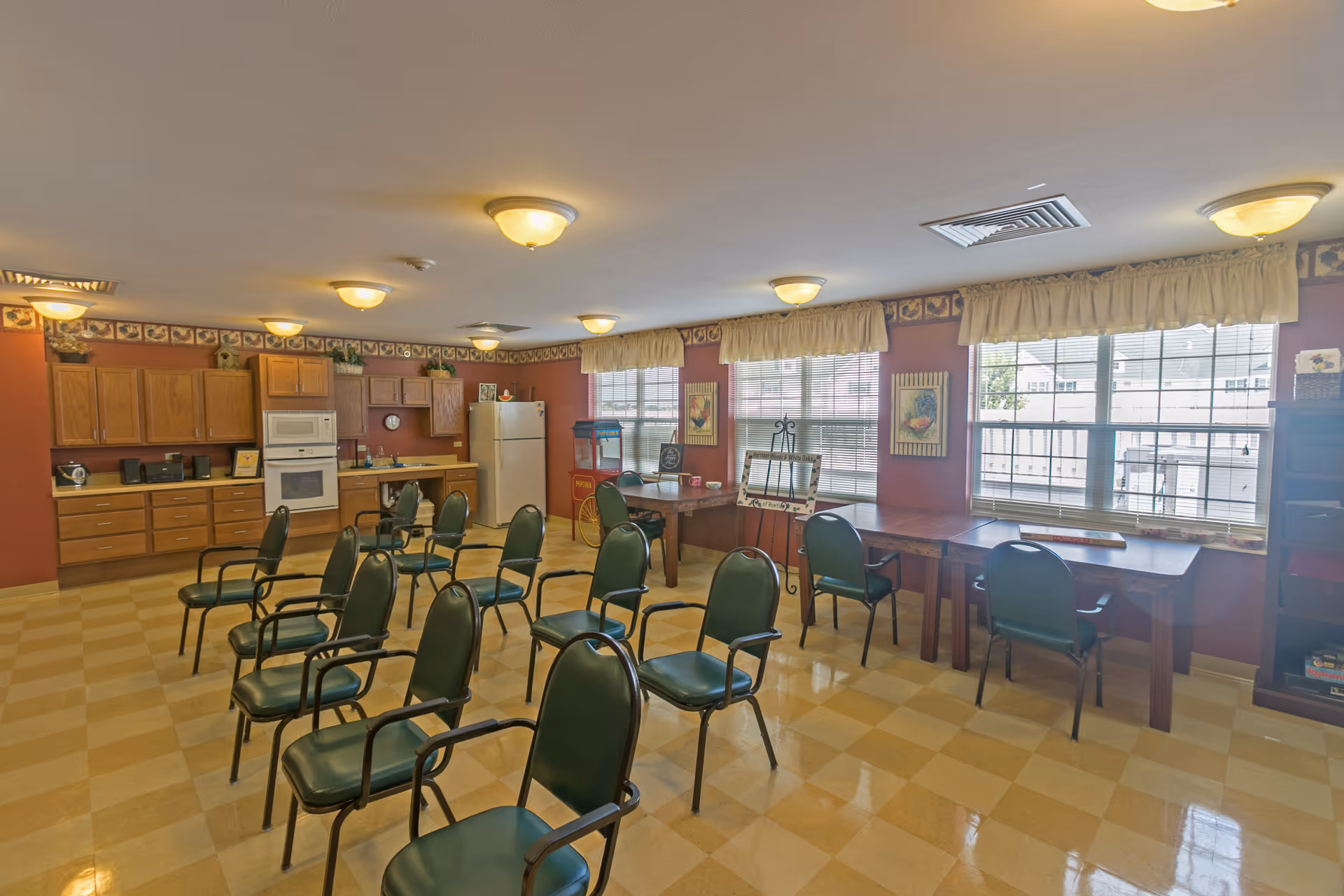 A spacious room with a kitchen area featuring wooden cabinets, a microwave, an oven, and a refrigerator. Several green chairs are arranged in rows facing the kitchen. The room has large windows with beige curtains, framed artwork on the walls, and a popcorn machine in the corner. The floor is checkered with yellow and beige tiles.