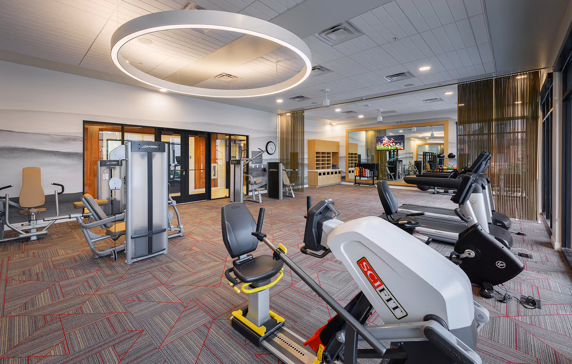 A spacious fitness room with various exercise equipment including a recumbent bike, treadmills, and weight machines. The room features a large circular ceiling light, a wall-mounted TV, a large mirror, and a storage area with cubbies. The floor is carpeted with a geometric pattern, and there are large windows with sheer curtains allowing natural light to enter.