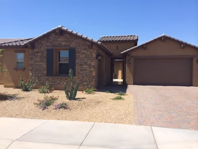 Front exterior view of a single-story house with stone and stucco facade, a tiled roof, a two-car garage, desert landscaping with gravel, a cactus, and some small plants under a clear blue sky.