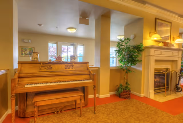 Wooden upright piano and bench beside a fireplace and potted plant in a cozy senior living common area.