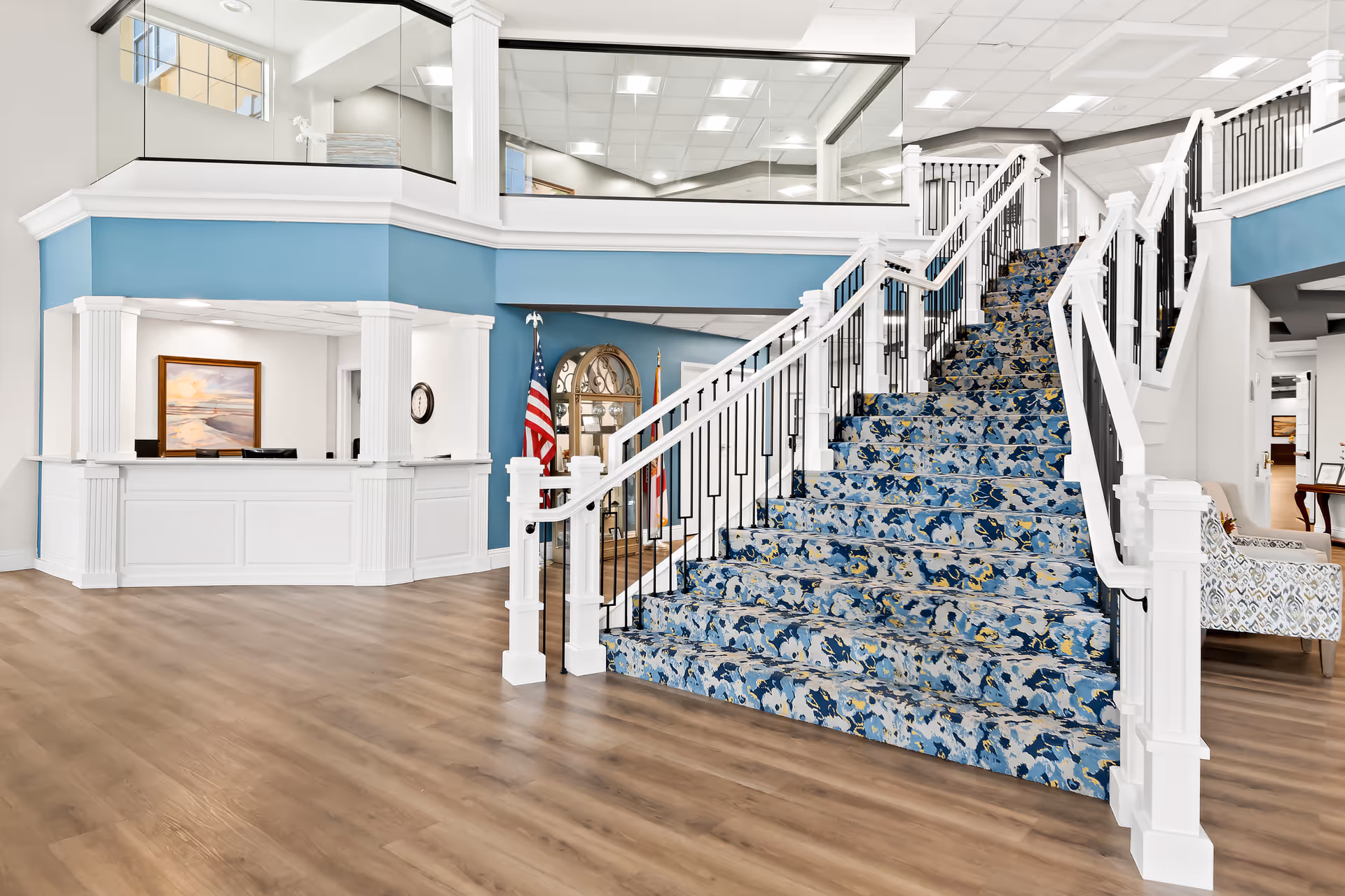 Bright and spacious interior of a senior living facility featuring a wide staircase with a blue patterned carpet, white railings, and wooden flooring. To the left is a reception desk with white paneling and blue walls behind it. An American flag and a clock are visible near the staircase. The area has large windows and a high ceiling with recessed lighting.
