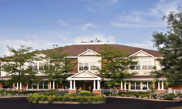 Front exterior view of a two-story senior living facility building with a red brick facade, white trim, and a covered entrance supported by white columns. The building is surrounded by landscaped greenery and trees under a partly cloudy sky.
