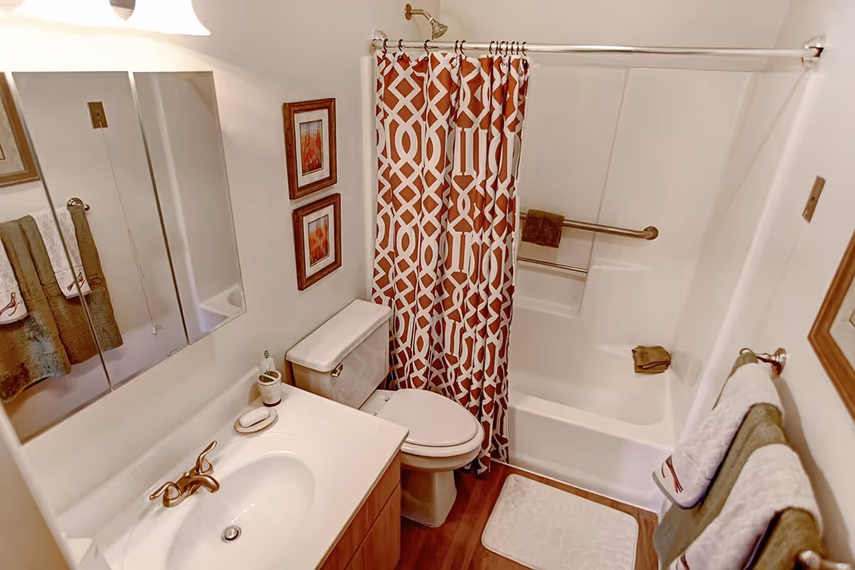 A clean bathroom featuring a white sink with a silver faucet, a toilet, and a bathtub with a red and white patterned shower curtain. The bathroom has wooden flooring, two framed pictures on the wall, and several towels hanging on towel racks.