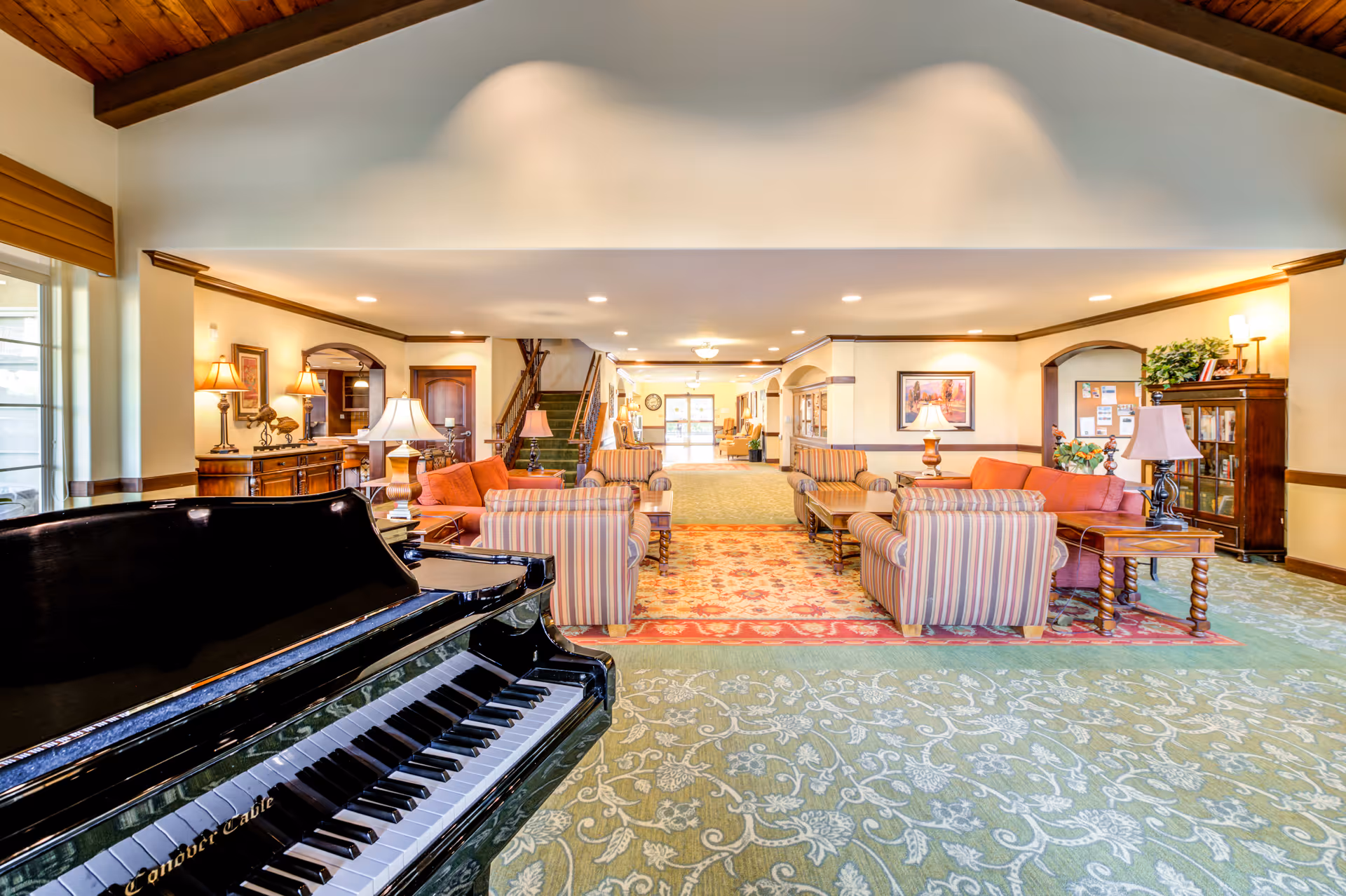 A spacious and well-lit senior living facility common area with a black grand piano in the foreground. The room features comfortable striped and solid orange sofas arranged around wooden coffee tables on a patterned carpet. The walls are adorned with framed artwork and lamps, and there is a staircase in the background leading to an upper floor. Large windows allow natural light to fill the space.