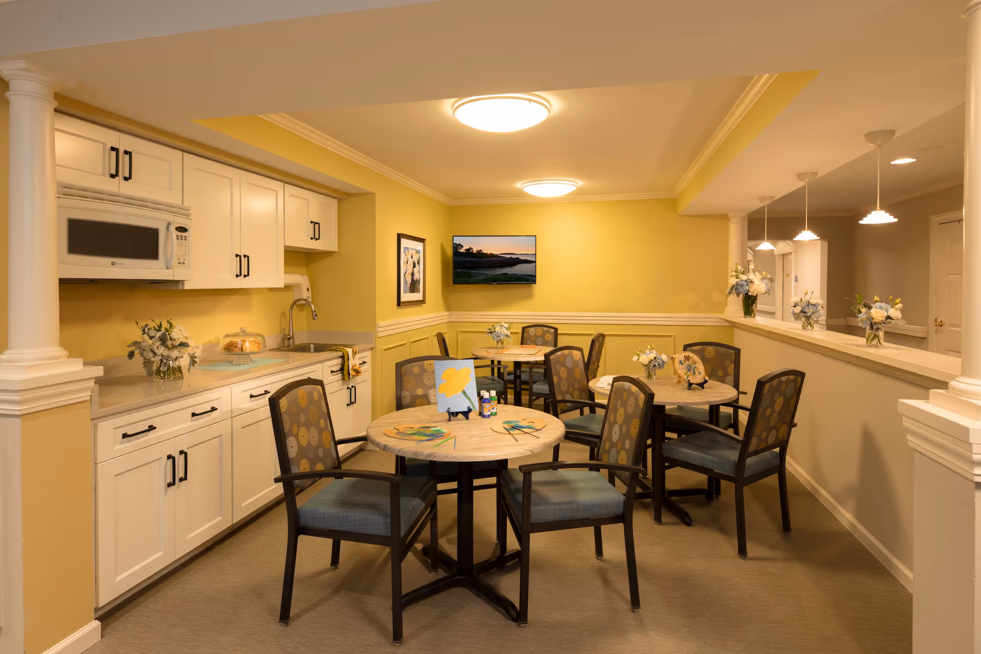 A cozy dining area with several round tables and chairs arranged around them. The walls are painted yellow and beige, and there are white cabinets with a microwave and sink along one side. The tables have small flower arrangements and some art supplies, including paintbrushes and a small canvas with a yellow flower painting. The room is softly lit with ceiling lights and pendant lights, and there is a TV mounted on the far wall displaying a nature scene.