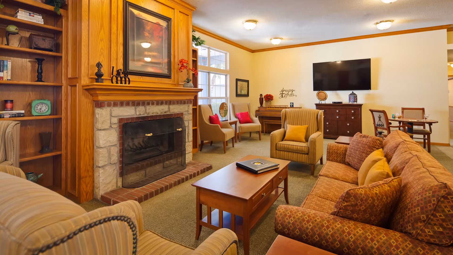 A cozy living room with a stone and wood fireplace, a wooden coffee table, a patterned sofa with multiple cushions, an armchair with a yellow pillow, and two beige chairs near a window. A flat-screen TV is mounted on the wall above a wooden cabinet, and a small dining table with chairs is visible in the background. The room is warmly lit with ceiling lights and decorated with framed artwork and small decorative items.
