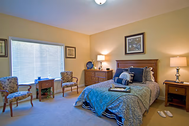 A cozy bedroom with a single bed covered in patterned blue and gray bedding, two wooden nightstands with lamps on either side, a wooden dresser with decorative items, two upholstered armchairs near a large window with blinds, and framed artwork on the walls.