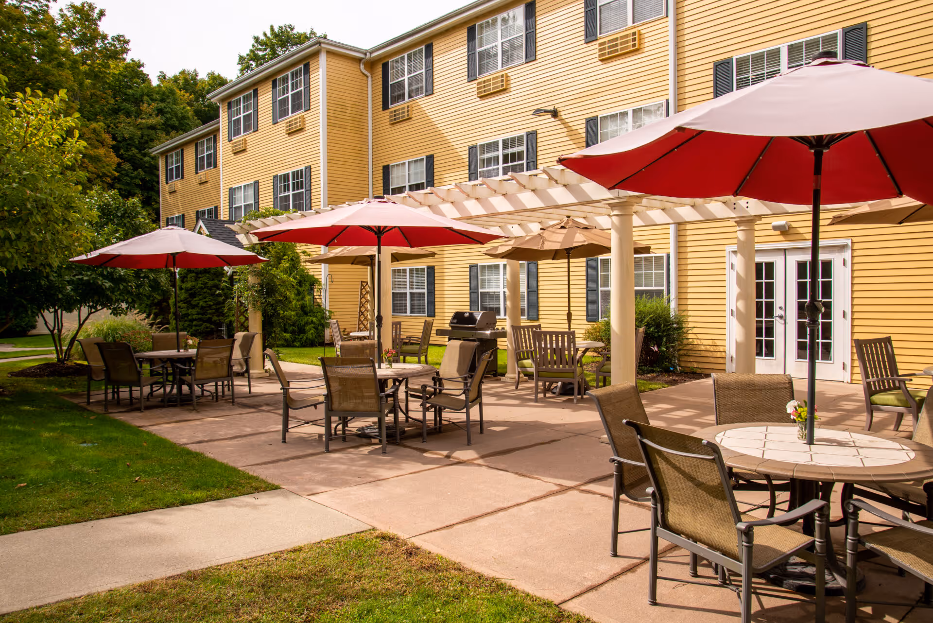 Sunlit outdoor patio with tables, chairs and red umbrellas in front of a yellow multi-story building.