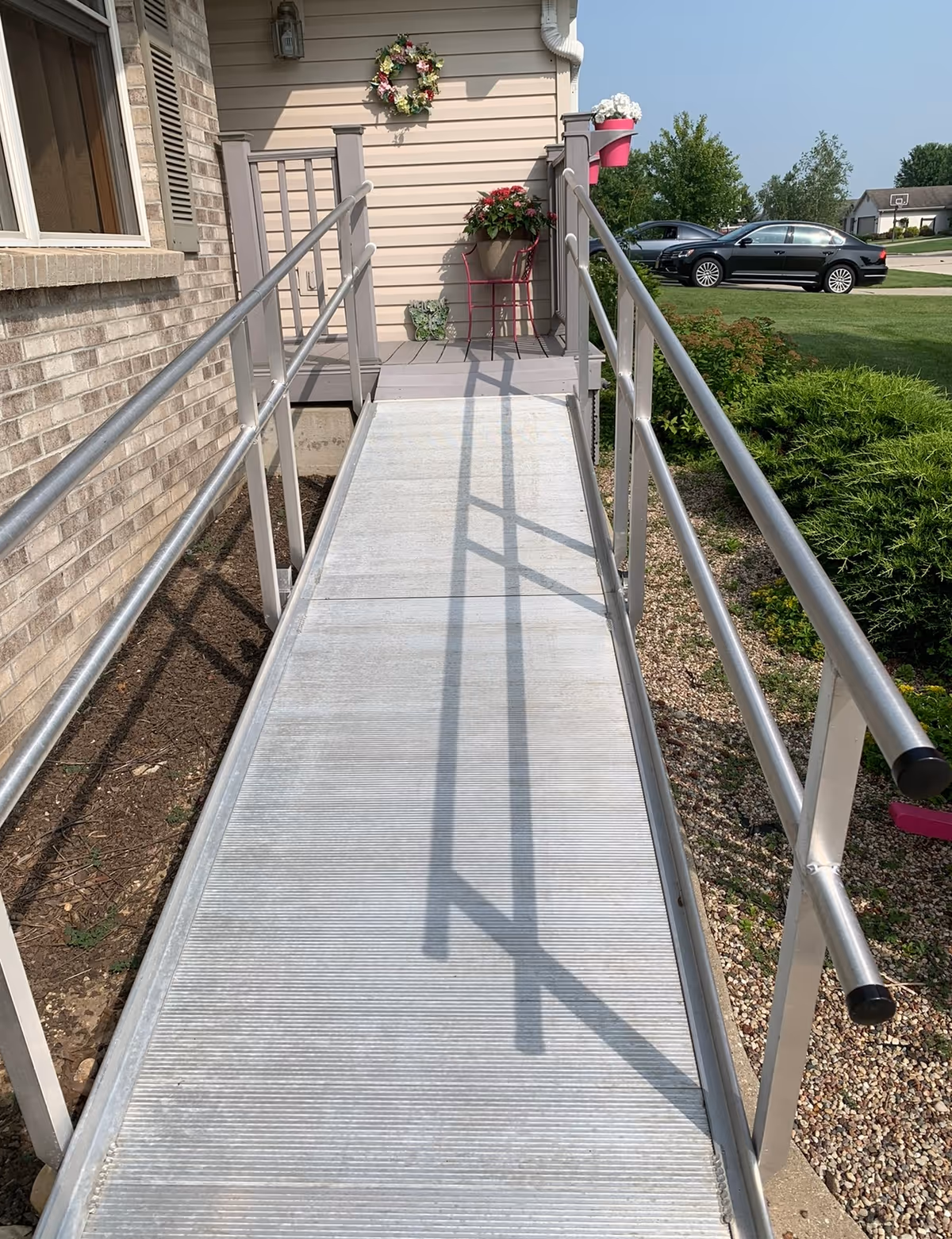 A metal wheelchair ramp leading up to a small porch area of a building. The porch has a flower wreath on the wall, a flower pot on a pink stand, and another flower pot attached to the railing. There is a car parked on the street and greenery around the ramp.