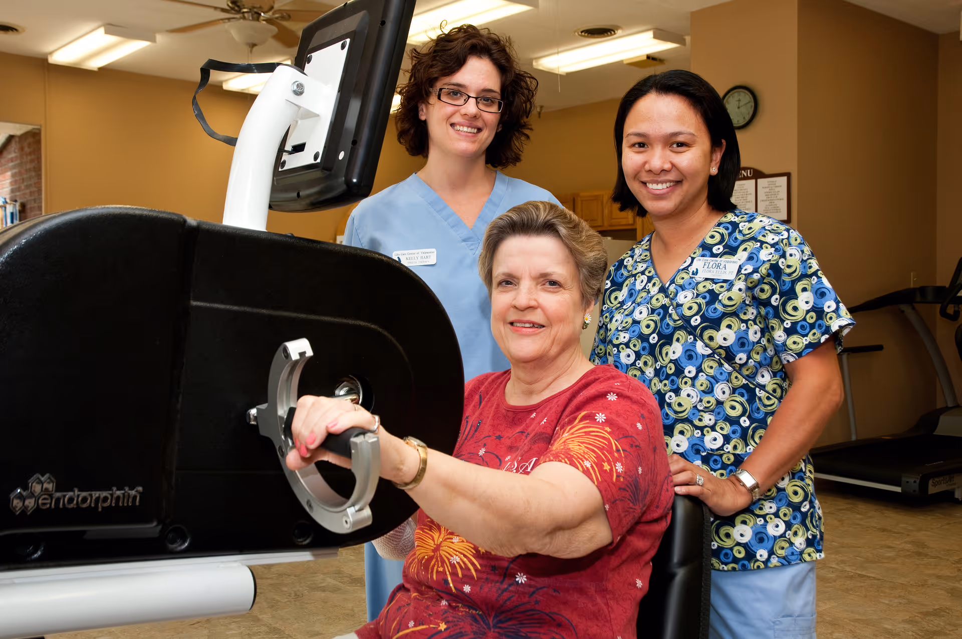 An elderly woman using a hand exercise machine in a fitness or rehabilitation room, accompanied by two female healthcare workers smiling behind her. The room has exercise equipment and a clock on the wall.