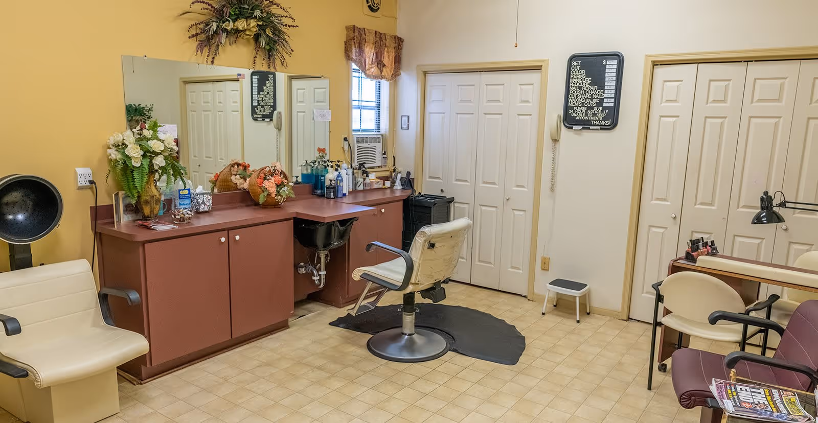 Interior salon area with a styling chair and sink at a counter, mirror, hair dryer, manicure station, and several waiting chairs.