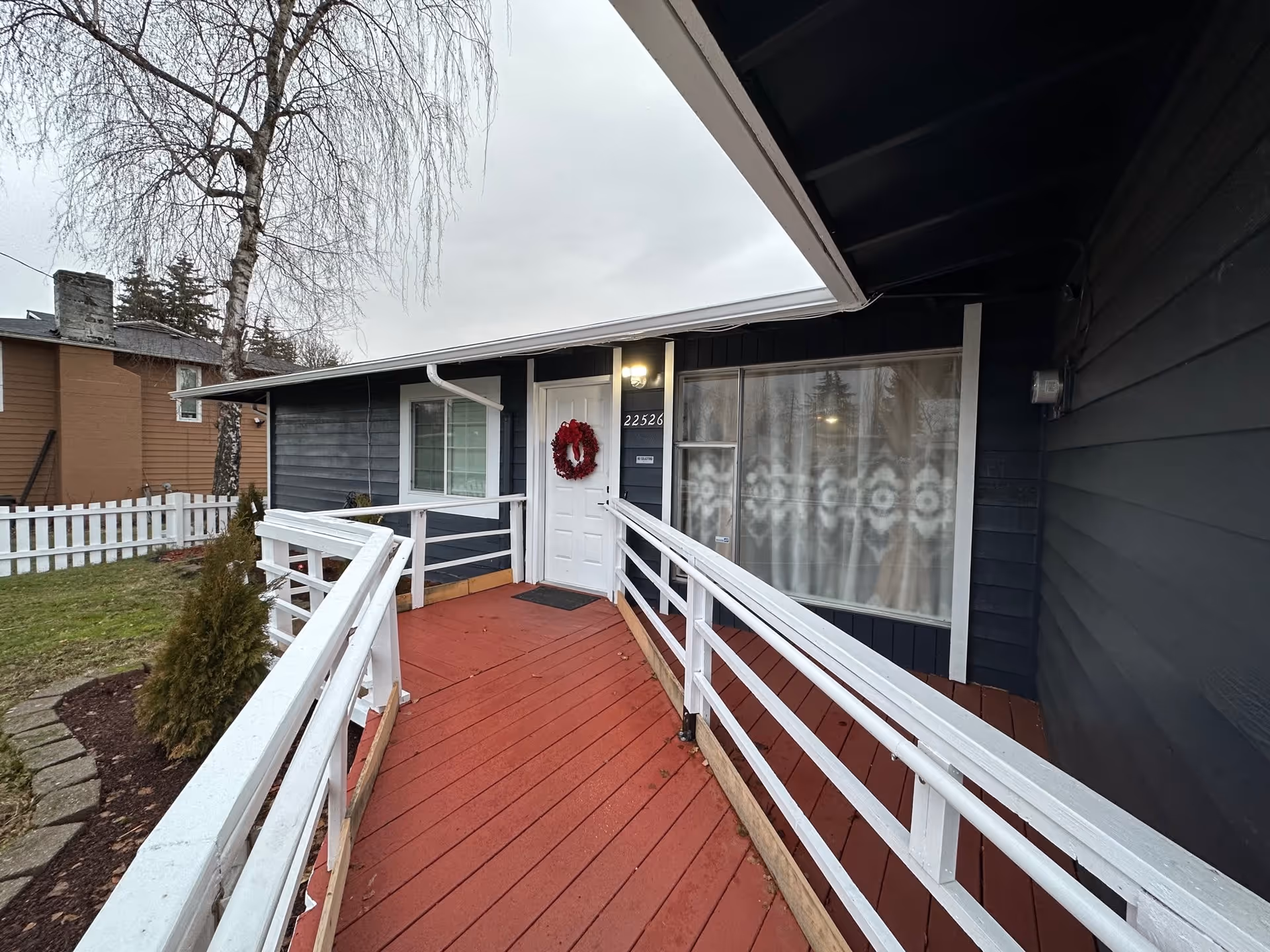 Exterior view of a single-story building with dark blue siding and white trim. A red wooden ramp with white railings leads to a white door decorated with a red wreath. The door is illuminated by a light above it. To the right of the door is a large window with patterned curtains. There is a small garden area with a tree and shrubs to the left, and a white picket fence in the background.