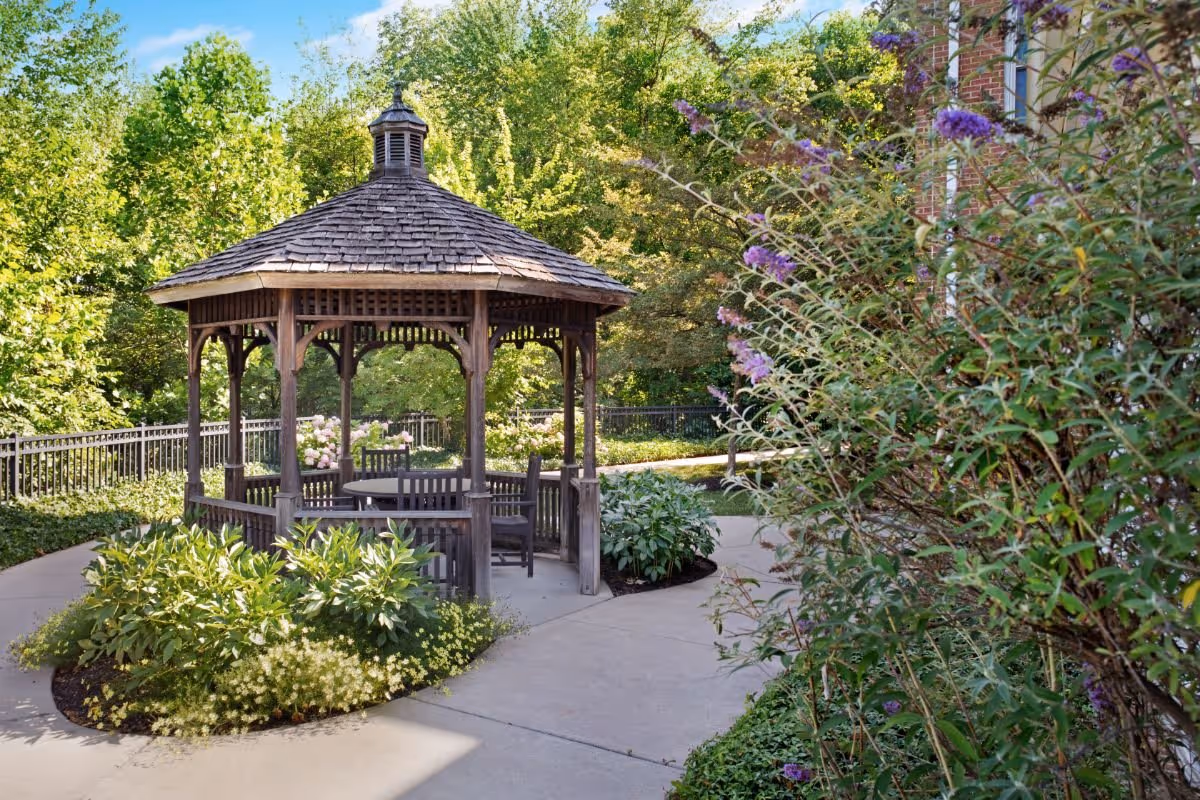A wooden gazebo with a shingled roof surrounded by lush green plants and flowers, situated on a paved pathway in a garden area with trees and shrubs in the background.
