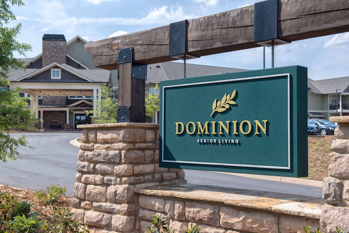 Stone and wood signpost with a green sign displaying 'DOMINION SENIOR LIVING' in gold letters, with a senior living facility building and driveway visible in the background under a partly cloudy sky.