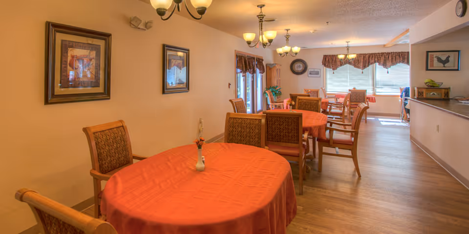 Interior view of a senior living facility dining room with several tables covered in orange tablecloths and wooden chairs with patterned cushions. The room is warmly lit with ceiling light fixtures and decorated with framed pictures on the walls and window valances. A small vase with flowers is placed on the nearest table.