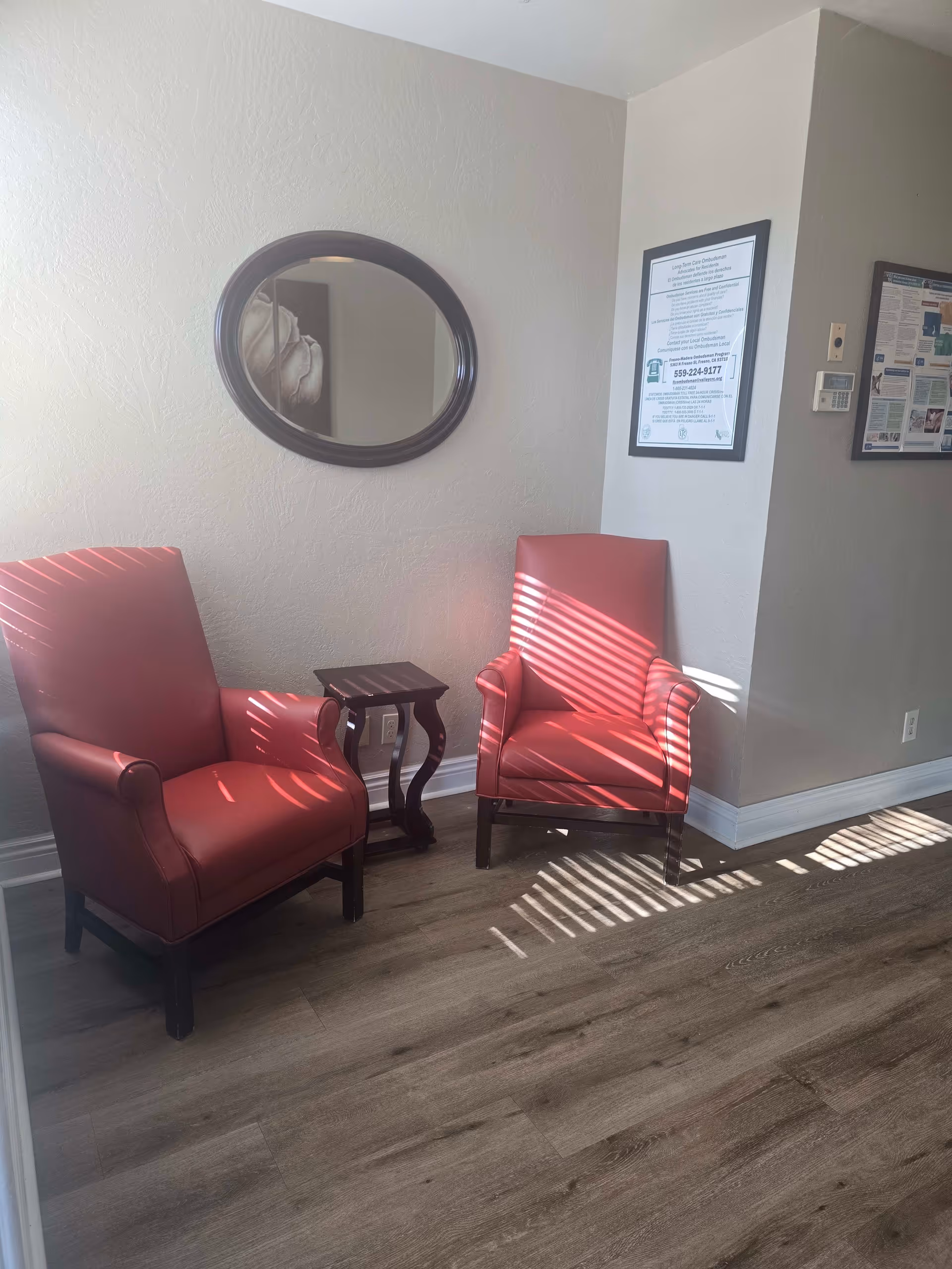 Two red armchairs with wooden legs positioned on either side of a small dark wooden side table in a room with light-colored walls and wood flooring. An oval mirror hangs on the wall above the left chair, and two framed notices are mounted on the adjacent wall. Sunlight streams through blinds, casting striped shadows on the chairs and floor.