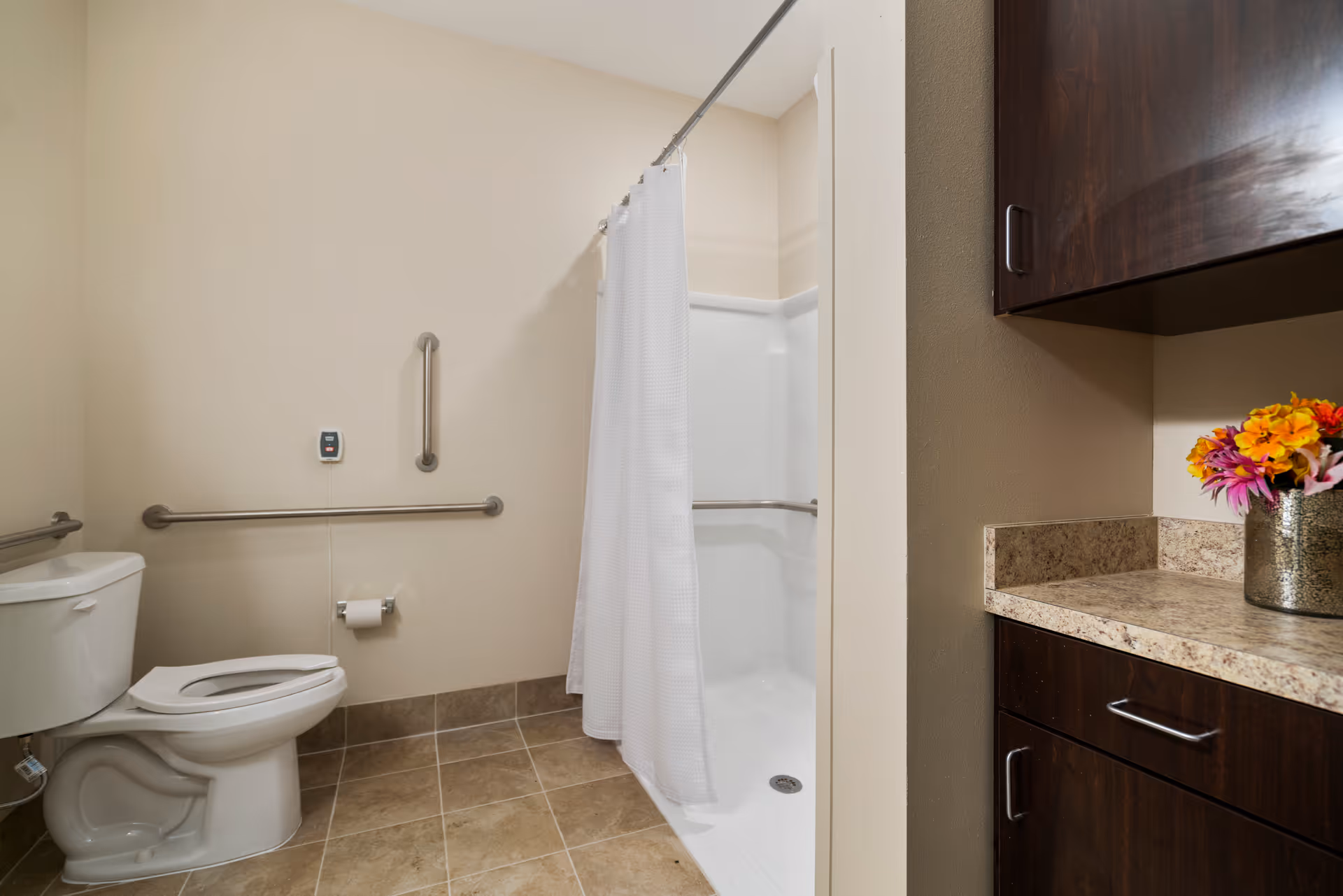 A clean and accessible bathroom featuring a white toilet with grab bars on the walls, a walk-in shower with a white curtain and grab bars, beige tiled floor, and dark wood cabinets with a countertop holding a vase of colorful flowers.