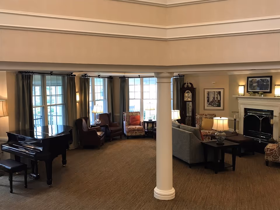 Spacious, warmly lit common living room with a grand piano, seating area around a fireplace, a central column, and large windows with curtains.