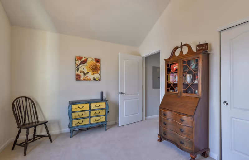 A room with beige walls and carpeted floor featuring a wooden chair on the left, a small blue and yellow chest of drawers with a floral painting above it, and a tall wooden cabinet with glass doors on the right. There are two white doors, one open and one closed.