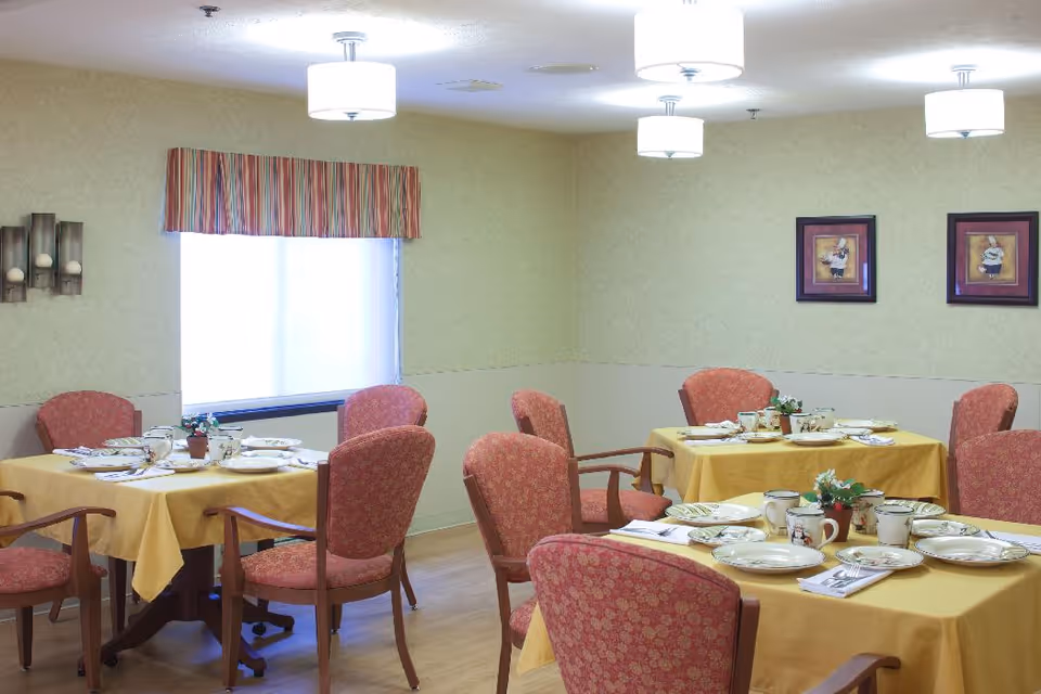 Dining room with several tables covered in yellow tablecloths, each set with plates, cups, and silverware. The room has pink upholstered chairs, light green walls with framed artwork, a window with a striped valance, and ceiling lights.