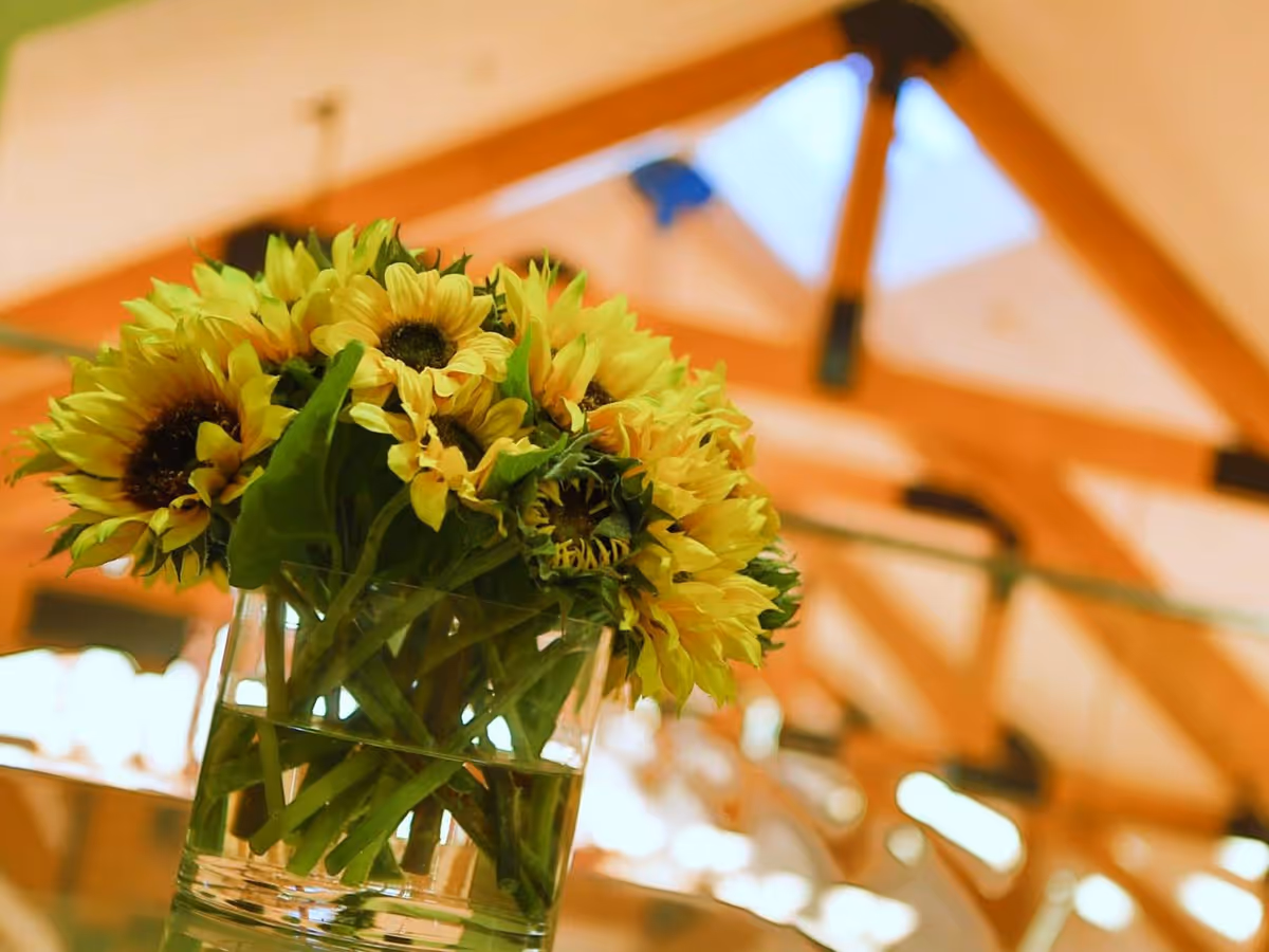 A close-up of a bouquet of yellow sunflowers in a clear glass vase filled with water, set indoors with a blurred background showing wooden beams and a skylight ceiling.