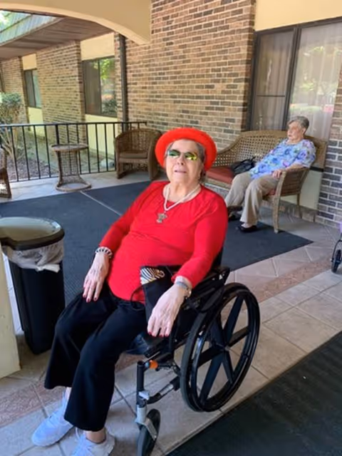 Two older women seated on a covered outdoor patio at a senior living facility, one in a wheelchair wearing a red hat and red top.