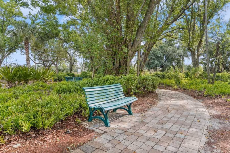 Green park bench next to a curved paved path surrounded by trees and landscaping.