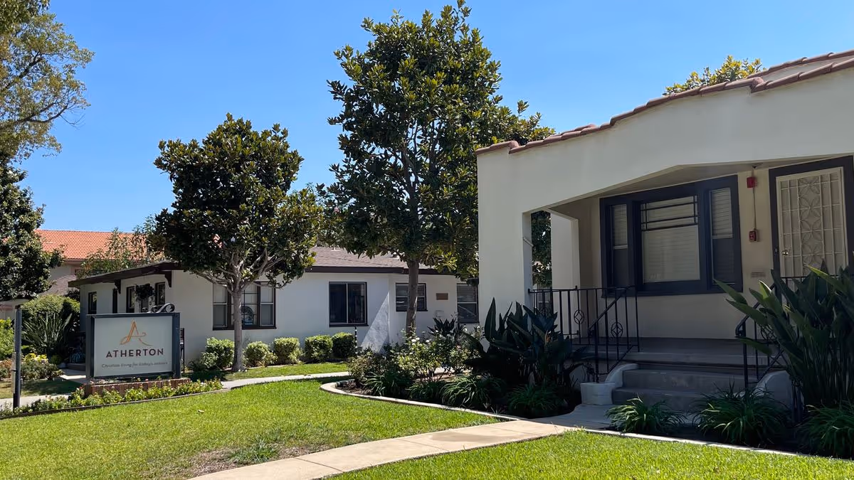 Exterior view of Atherton Baptist Homes with a manicured lawn, trees, a pathway, a sign, and the front porches of low-rise buildings.