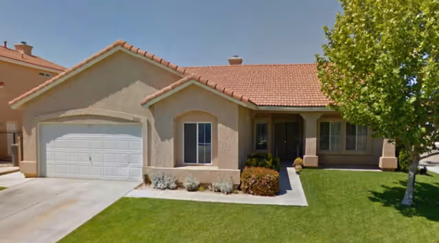 A single-story beige stucco building with a two-car garage, front yard, and tree.