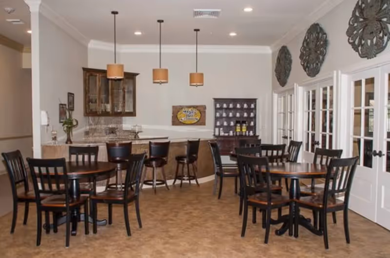 A dining area with several round wooden tables and black chairs. There is a counter with bar stools and pendant lights hanging above it. The walls are decorated with three large ornamental metal wall hangings and a wooden cabinet with cups and bottles. French doors are visible on the right side.