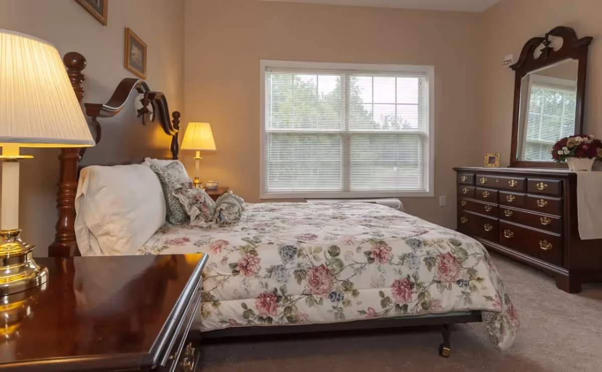 Traditional bedroom featuring a floral-covered bed with a wooden headboard, matching dresser, bedside lamps, and a window.