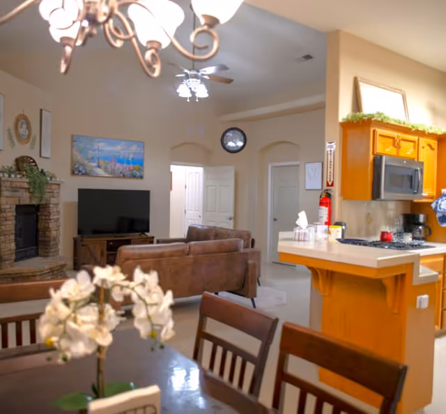 Open-plan interior with a dining table and flowers in the foreground, a living room with couch, TV and fireplace, and a kitchen island with cabinets.