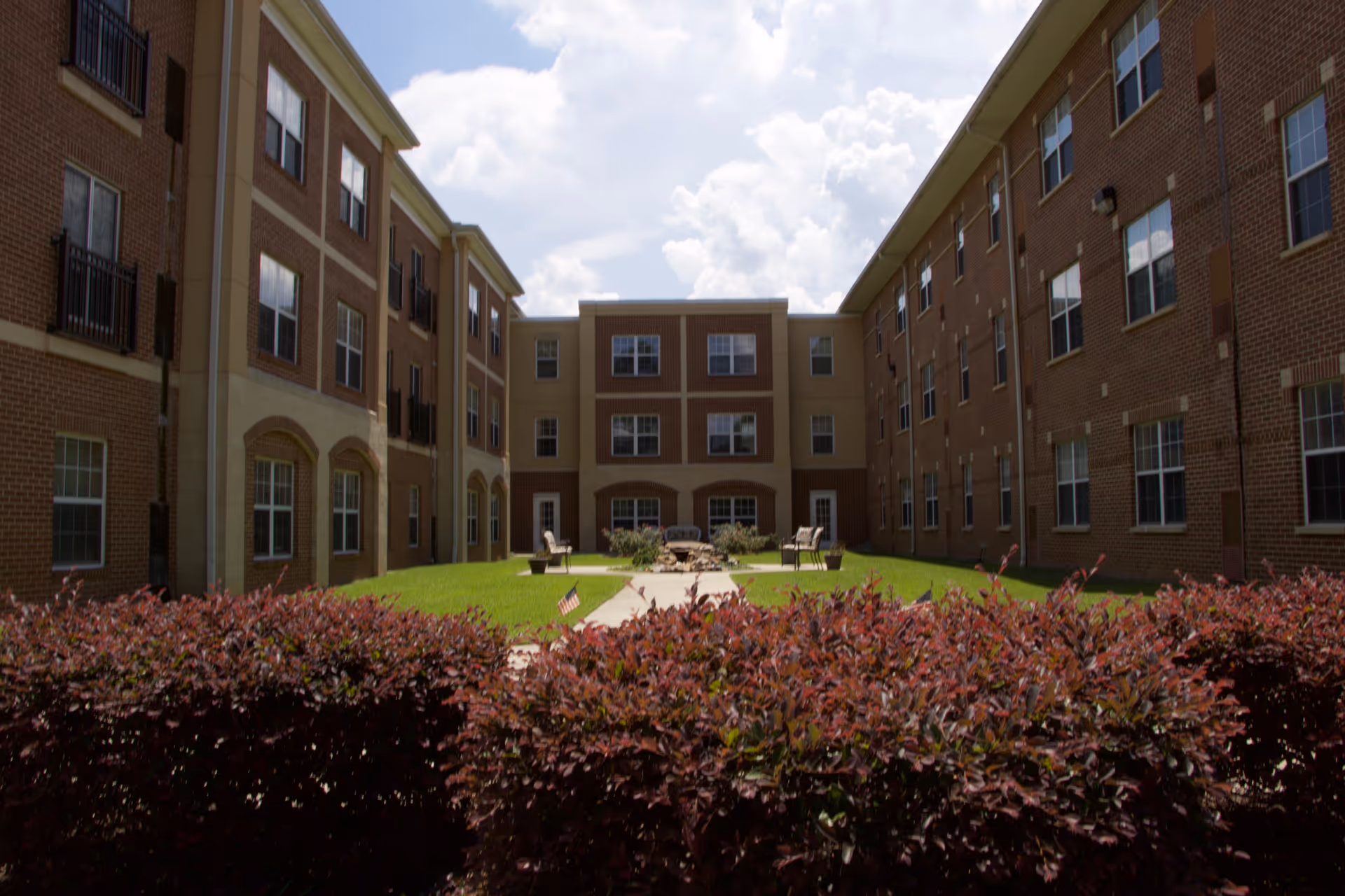 Outdoor courtyard area between two three-story brick buildings with a green lawn, a paved walkway, chairs, and a fire pit under a partly cloudy sky.