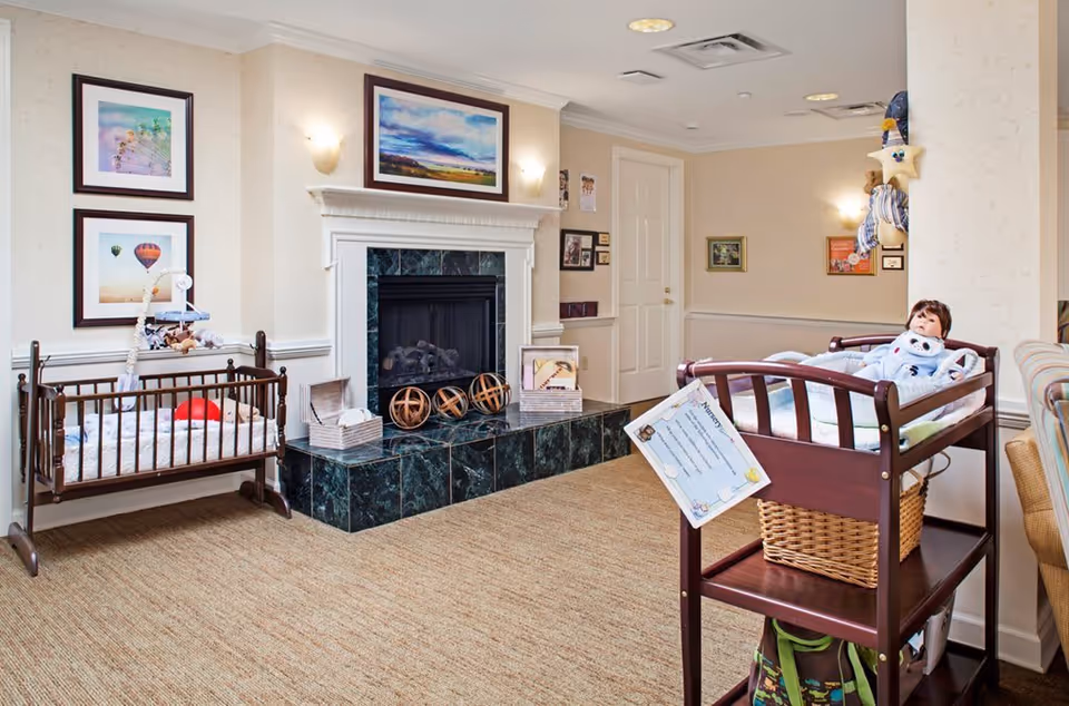 A cozy nursery room with a wooden crib containing a red ball and a mobile, a fireplace with decorative items on the hearth, framed pictures on the wall, and a changing table with a doll dressed in blue pajamas on top. The room has beige walls, carpeted floor, and soft lighting.