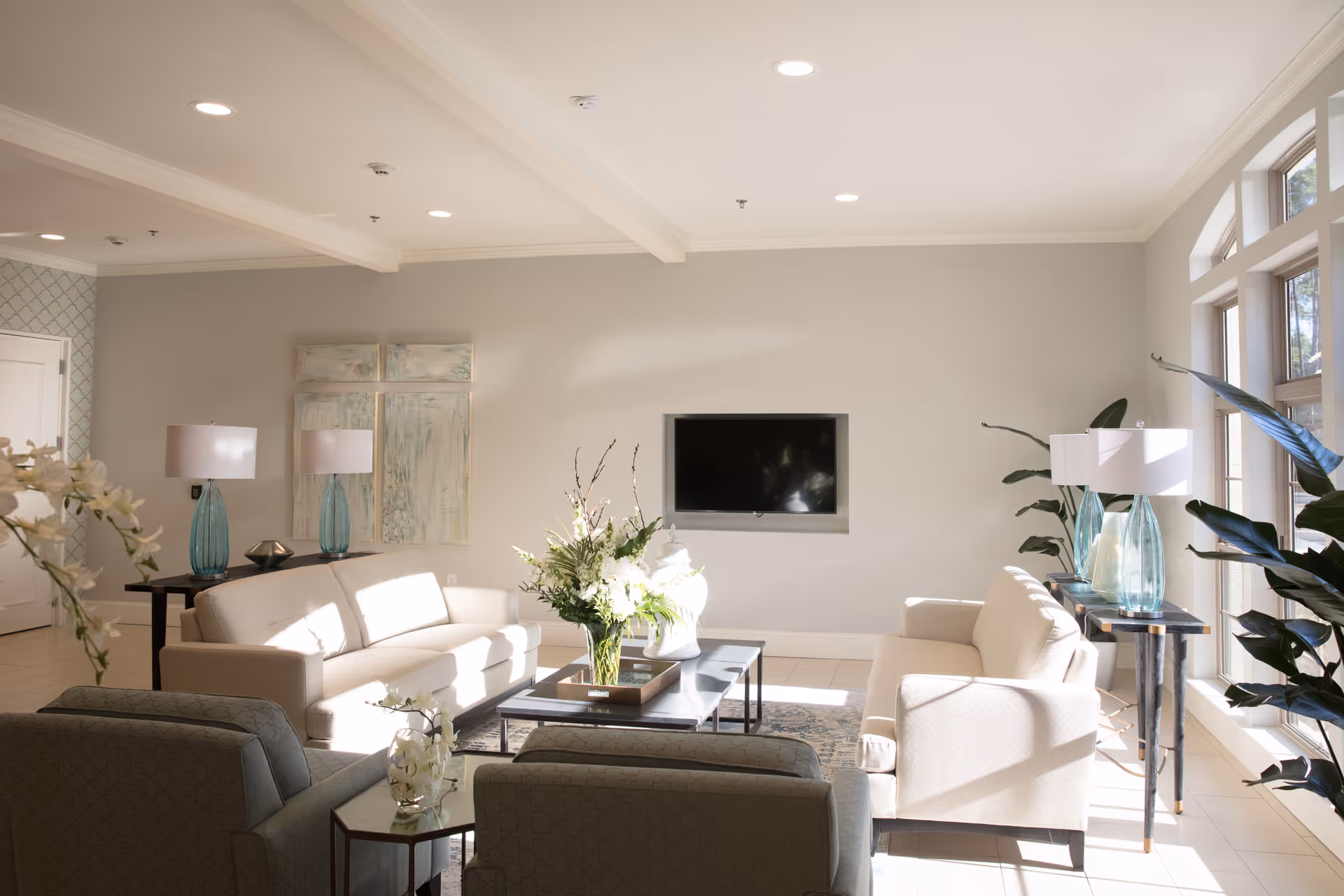 A bright and airy living room with large windows letting in natural light. The room features two beige sofas, two armchairs, a coffee table with a floral arrangement, side tables with blue glass lamps, and a wall-mounted flat-screen TV. There are decorative plants and artwork on the walls, creating a comfortable and inviting atmosphere.