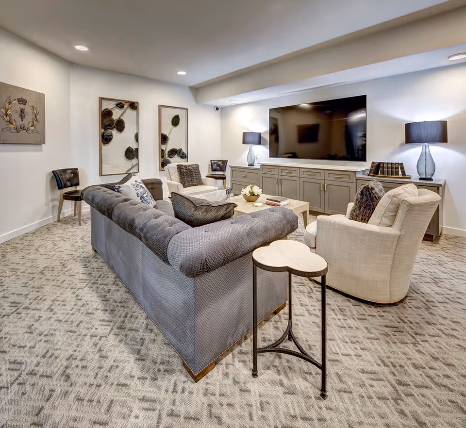 A cozy living room with a large gray sofa, two beige armchairs, a wooden coffee table with books and a flower arrangement, and a large flat-screen TV mounted on a gray cabinet. The room has patterned carpet, two table lamps with dark shades on either side of the TV, two framed botanical prints on the wall, and a small side table next to the sofa.