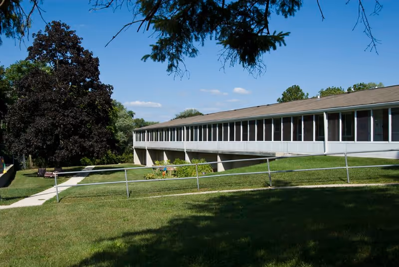 A long single-story building with a covered porch or walkway supported by columns, surrounded by green grass and trees under a clear blue sky.