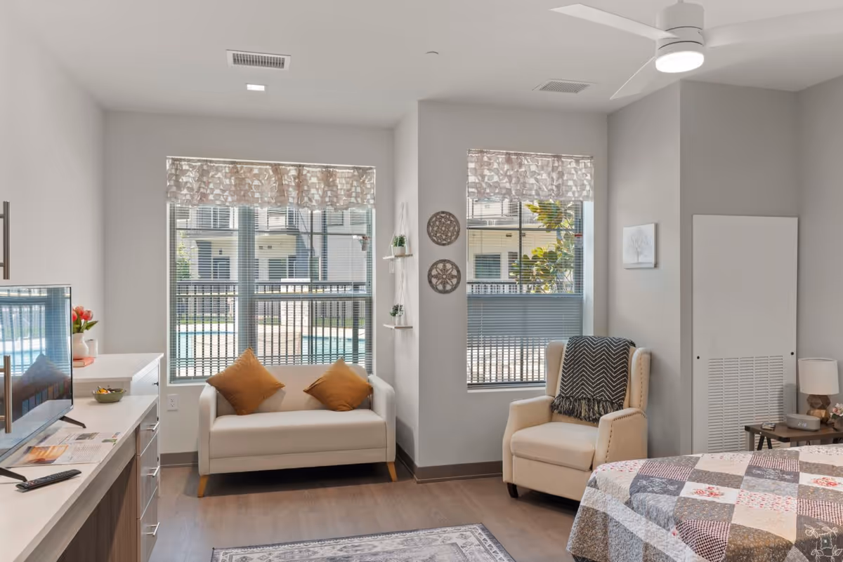 Bright senior studio room with a small couch, armchair, TV, and a corner of a bed by two large windows.