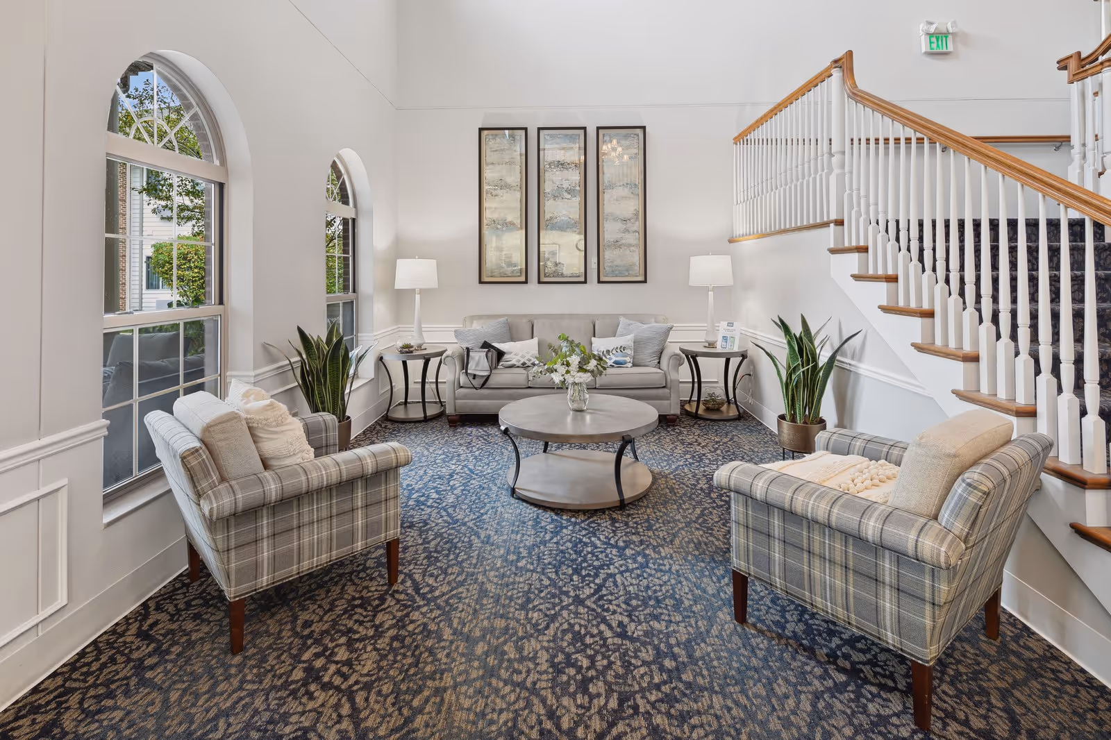Bright communal seating area with upholstered chairs and a sofa around a round coffee table beside arched windows and a staircase.