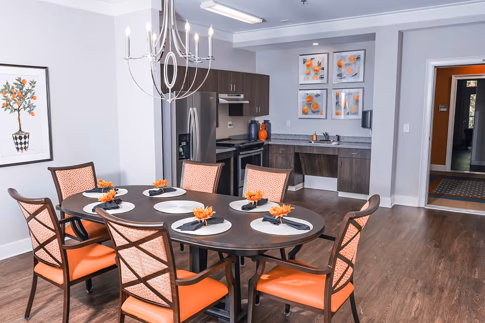 Dining area with an oval table set for six and orange-upholstered chairs, with a kitchenette and wall art in the background.
